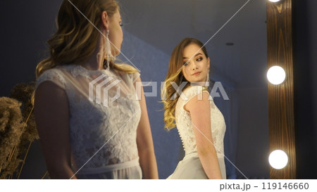 Beautiful bride with long hair is wearing a white lace wedding dress and looking in a mirror Beautiful bride with long hair is wearing a white lace wedding dress and looking in a mirror 119146660