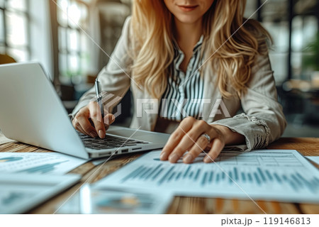 Office worker woman is sitting at a desk with a laptop and a stack of papers and writing on the papers with a pen, working on a project or a report 119146813