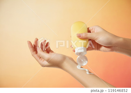 The minimalist composition image features a model hand squeezing a yellow tube with a white cap off a cream piece, a bright orange background resembling the sun, suitable for sunscreen commercials. 119146852