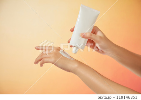 The female model gently squeezes a sample of cream onto her other hand from an unlabeled white tube under a bright orange background, suitable for a sunscreen advertisement. 119146853