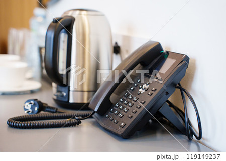 A corded black telephone in the hotel room on a table with a kettle and cups A corded black telephone in the hotel room on a table with a kettle and cups 119149237