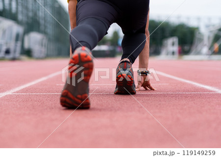 Close-up view of a runner's legs, primed in a low start position, signaling the beginning of an energetic sprint along the urban outdoor stadium's red rubberized track.  119149259