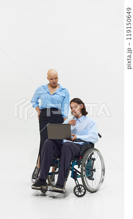 Young bald woman in blue shirt standing next to man in wheelchair working on laptop, showcasing inclusivity and collaboration in workplace. 119150649