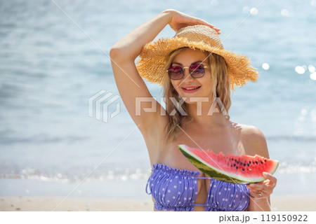 Portrait of young beautiful smiling blond woman in swimsuit wearing sunglasses and hat while happily sitting with slice of watermelon on beach Portrait of young beautiful smiling blond woman in swimsuit wearing sunglasses and hat while happily sitting with slice of watermelon on beach 119150922