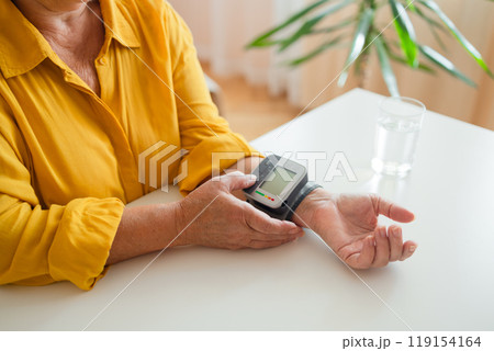 Senior woman with hypertension measuring blood pressure herself at home. Glass of water on the table Senior woman with hypertension measuring blood pressure herself at home. Glass of water on the table 119154164