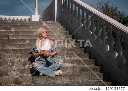 A woman sits on a set of stairs, holding a cup of coffee. She is enjoying her coffee and taking in the view from the top of the stairs. 119155777