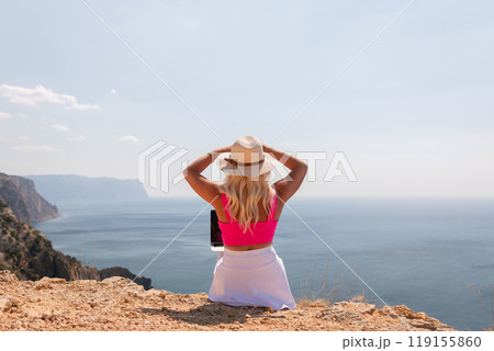 A woman in a pink top and white skirt is sitting on a rocky hill overlooking the ocean. She is wearing a straw hat and has a laptop open in front of her. Concept of relaxation and leisure. A woman in a pink top and white skirt is sitting on a rocky hill overlooking the ocean. She is wearing a straw hat and has a laptop open in front of her. Concept of relaxation and leisure. 119155860