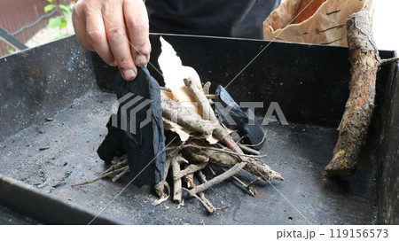 Hands place a piece of light brown paper among small sticks and twigs varying in color from beige to dark brown, which are placed on a black metal tray or fire pit. Hands place a piece of light brown paper among small sticks and twigs varying in color from beige to dark brown, which are placed on a black metal tray or fire pit. 119156573