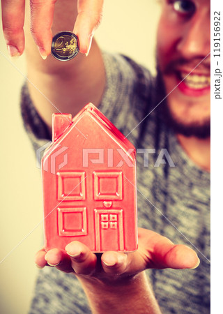 Smiling man putting coin into house piggybank Smiling man putting coin into house piggybank 119156922