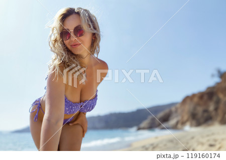 A beautiful girl in sunglasses on the beach looks at the camera, leaning towards it. Sexy blonde in a swimsuit on the seashore 119160174