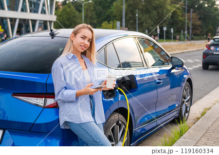 Blonde woman, wearing casual clothes stands beside a charging electric car, sipping coffee and checking her smartphone 119160334