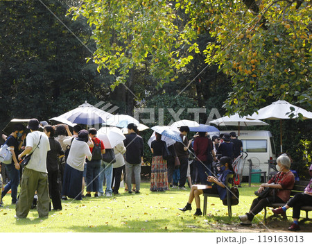 公園のイベント会場に並んだキッチンカー。日傘をさしながら綺麗に並んだ人々。柿の実の下の日陰で休む人も 119163013