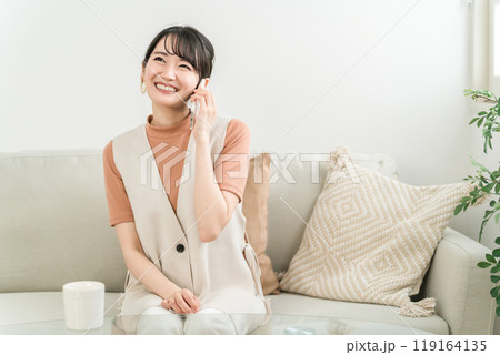 ソファに座ってスマホで友達と電話する笑顔のアジア人女性 119164135