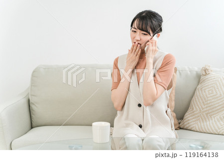 スマホで電話・通話・会話しながら驚く・びっくりするソファに座ったアジア人女性 119164138