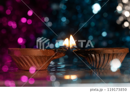 Two illuminated traditional clay diyas with glowing flames on a vibrant bokeh background of colorful pink and blue colors. 119173850