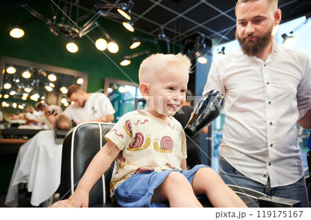 Cheerful child in barbershop chair at the end of haircut, barber using hairdryer to remove loose hair clippings. Enjoyment or satisfaction with the haircut experience. Cheerful child in barbershop chair at the end of haircut, barber using hairdryer to remove loose hair clippings. Enjoyment or satisfaction with the haircut experience. 119175167