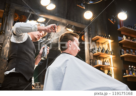 Professional barber using hairspray to finish hairstyle. Stylish hairdresser making final touches. Young customer getting fresh haircut. Professional barber using hairspray to finish hairstyle. Stylish hairdresser making final touches. Young customer getting fresh haircut. 119175168