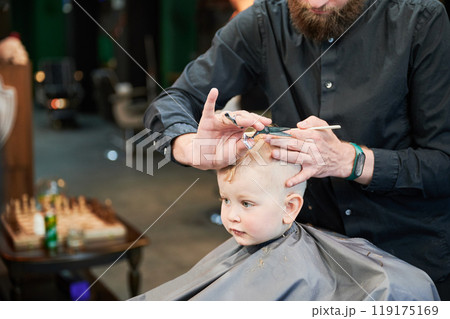Man barber cutting little boy's hair using comb and scissors. Child getting haircut from adult male, likely barber. Professional hairdresser and cute client at modern barbershop. Man barber cutting little boy's hair using comb and scissors. Child getting haircut from adult male, likely barber. Professional hairdresser and cute client at modern barbershop. 119175169