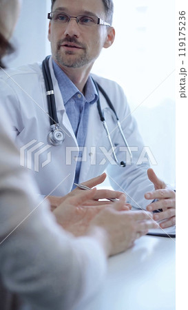 Doctor and a patient. The physician, wearing a white medical coat over a blue shirt, gesturing with his hands during a consultation in the clinic. Medicine concept Doctor and a patient. The physician, wearing a white medical coat over a blue shirt, gesturing with his hands during a consultation in the clinic. Medicine concept 119175236