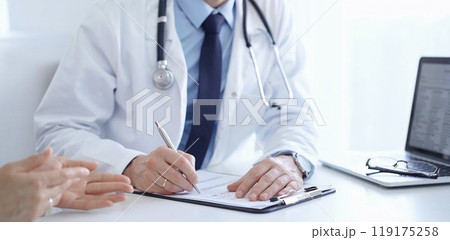A doctor and a patient. The physician, wearing a white medical coat over a blue shirt and tie, is filling out a medical record form during a consultation in the clinic. Medical service 119175258