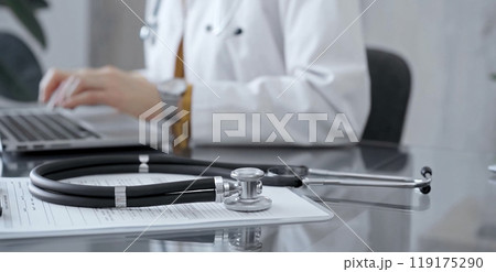 Stethoscope and clipboard with medication history records are on a glass desk behind a physician using a laptop computer. Medicine concept Stethoscope and clipboard with medication history records are on a glass desk behind a physician using a laptop computer. Medicine concept 119175290