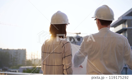 Two architects, wearing safety helmets and glasses, are examining blueprints at a construction site during sunrise or sunset, viewed from the back. Architecture and engineering concepts Two architects, wearing safety helmets and glasses, are examining blueprints at a construction site during sunrise or sunset, viewed from the back. Architecture and engineering concepts 119175292