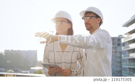 Two architects colleagues, wearing safety helmets and glasses, are standing near each other and discussing a construction site progress during sunrise or sunset. Architecture and engineering Two architects colleagues, wearing safety helmets and glasses, are standing near each other and discussing a construction site progress during sunrise or sunset. Architecture and engineering 119175305