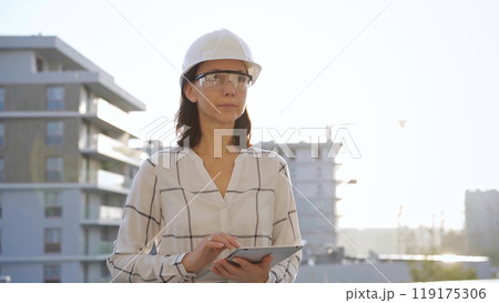 Woman construction engineer wearing white checked blouse and hard hat is using tablet computer while inspecting a building site at sunset, front view. Architecture and engineering concepts Woman construction engineer wearing white checked blouse and hard hat is using tablet computer while inspecting a building site at sunset, front view. Architecture and engineering concepts 119175306