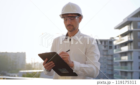 Man construction engineer wearing white shirt and hard hat is making notes on a clipboard while inspecting a building site at sunset, front view. Architecture and engineering concepts Man construction engineer wearing white shirt and hard hat is making notes on a clipboard while inspecting a building site at sunset, front view. Architecture and engineering concepts 119175311
