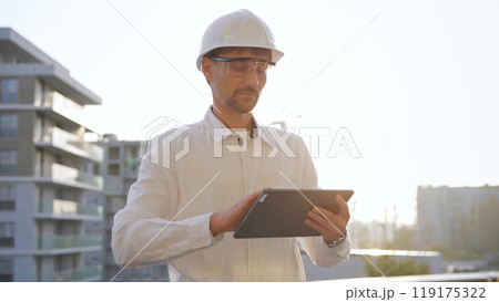 Man construction engineer or architect wearing white shirt and hard hat is using digital tablet while inspecting a building site at sunset, front view. Architecture and engineering concepts 119175322