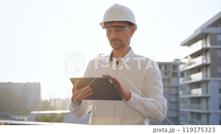Man construction engineer or architect wearing white shirt and hard hat is using digital tablet while inspecting a building site at sunset, front view. Architecture and engineering concepts 119175323