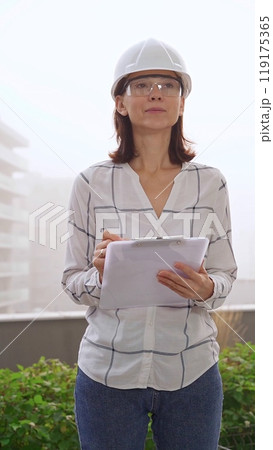 Female construction engineer wearing white checked blouse and safety helmet is making notes on a clipboard while inspecting a building site in foggy weather, front vertical view. Architecture concept 119175365