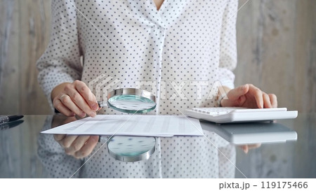 Auditor woman analyzing financial documents with magnifying glass and calculator at her office desk in formal white blouse with polka dots. Business people concept Auditor woman analyzing financial documents with magnifying glass and calculator at her office desk in formal white blouse with polka dots. Business people concept 119175466