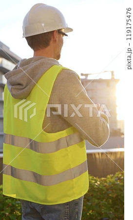 Man an architect or engineer with white helmet and safety vest is taking notes on a digital tablet while inspecting a construction site at sunrise, vertical back view. Architecture concept Man an architect or engineer with white helmet and safety vest is taking notes on a digital tablet while inspecting a construction site at sunrise, vertical back view. Architecture concept 119175476