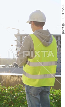 Man engineer with white helmet and safety vest is taking notes on a clipboard while inspecting a construction site at sunrise, back vertical view 119175500