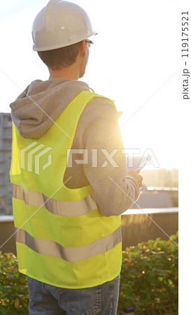 Man an architect or engineer with white helmet and safety vest is taking notes on a digital tablet while inspecting a construction site at sunrise, vertical back view. Architecture concept Man an architect or engineer with white helmet and safety vest is taking notes on a digital tablet while inspecting a construction site at sunrise, vertical back view. Architecture concept 119175521