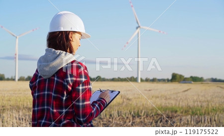 Female engineer wearing a white protective helmet is taking notes with a clipboard in a field with wind turbines, as the sun sets. Clean energy concept Female engineer wearing a white protective helmet is taking notes with a clipboard in a field with wind turbines, as the sun sets. Clean energy concept 119175522