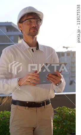 Male construction engineer wearing white shirt and helmet is using digital tablet while inspecting a building site at sunrise, front and vertical view . Architecture concept 119175538