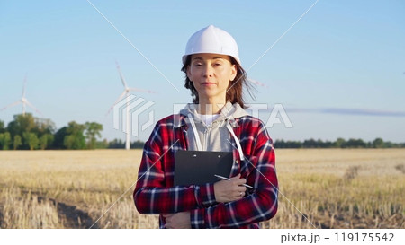 Woman engineer wearing white protective helmet is standing straight in a field with wind turbines, as the sun sets. Concept of clean energy and engineering audit 119175542