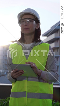 Woman a construction engineer with a white hard hat and safety vest is using digital tablet while inspecting a construction site in the early morning at sunrise, front view Woman a construction engineer with a white hard hat and safety vest is using digital tablet while inspecting a construction site in the early morning at sunrise, front view 119175546