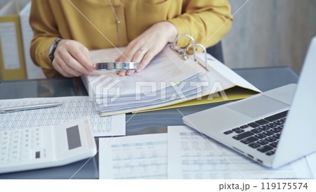 Female auditor in business attire inspecting financial reports with magnifying glass, surrounded by documents and a laptop and calculator. Business audit and taxes concept 119175574