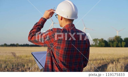 Man engineer at work is wearing a white protective helmet and taking notes with a clipboard in a field with wind turbines, as the sun sets. Clean energy concept 119175833