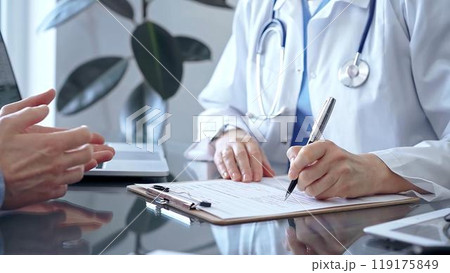 Doctor woman wearing white medical coat is consulting patient in clinic office while both are sitting at the glass desk. Physician is listening and making notes. Medicine concept Doctor woman wearing white medical coat is consulting patient in clinic office while both are sitting at the glass desk. Physician is listening and making notes. Medicine concept 119175849