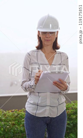 Woman a construction engineer wearing white checked blouse and hard hat is making notes on a clipboard while inspecting a building site in foggy weather, front vertical view. Architecture concept 119175851