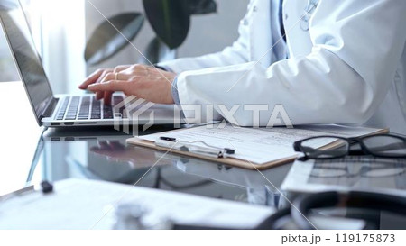 Doctor man with blue shirt and medical coat is using laptop computer in clinic office while sitting at the glass desk. Medicine concept 119175873