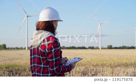 Female engineer wearing a white protective helmet is taking notes with a clipboard in a field with wind turbines, as the sun sets. Clean energy concept Female engineer wearing a white protective helmet is taking notes with a clipboard in a field with wind turbines, as the sun sets. Clean energy concept 119175878
