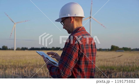 Man engineer at work is taking notes with a tablet computer in a field with wind turbines, as the sun sets. Clean energy concept Man engineer at work is taking notes with a tablet computer in a field with wind turbines, as the sun sets. Clean energy concept 119175882