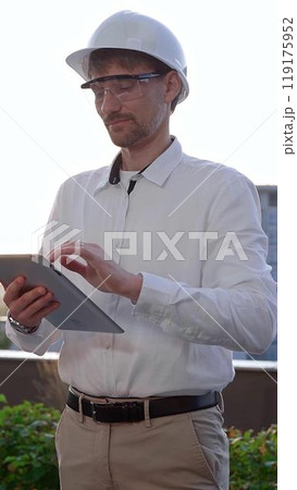 Man an architectural engineer, wearing a white shirt and hard hat, is using a digital tablet while inspecting a building site at sunrise, front and vertical view 119175952