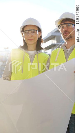 Two architects wearing safety hard hats and vests holding blueprint and discussing something on construction site at sunrise, front vertical view. Architecture and engineering concepts 119175999