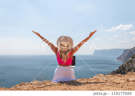 A woman is sitting on a rock by the ocean with a laptop open in front of her. She is wearing a pink top and a straw hat. Concept of relaxation and leisure. 119177089
